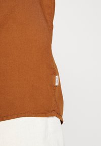 Top in tessuto marrone con texture, dotato di una cucitura laterale visibile e un piccolo etichetta bianca del marchio al bordo.