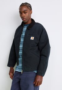 Young man with braided hair wears a black Carhartt jacket over a blue striped shirt and navy pants, standing against a white background.