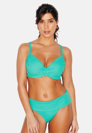 Donna che indossa un bikini turchese con dettaglio ad anello sulla parte superiore e slip a vita alta, rivolta in avanti su uno sfondo semplice.