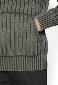 Main dans la poche d'un pull en maille côtelée verte avec des motifs épais verticaux et horizontaux, associé à un pantalon noir.