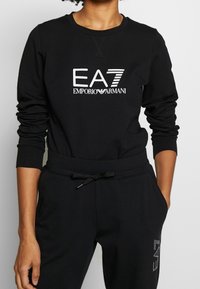 Svart sweatshirt och byxor med "EA7 EMPORIO ARMANI"-logotyp i vitt, mjukt tyg, avslappnad passform, ribbade ärmslut och dragsko i midjan.
