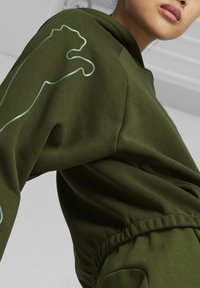 Olijfgroene hoodie met een losse pasvorm, voorzien van een witte omtrek van een pootafdruk op de linkermouw en een textuurvol stof. Trekkoord aan de capuchon.