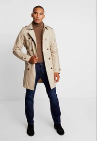 Abrigo tipo gabardina beige con botones dobles, hombreras y un ajuste entallado, llevado sobre un jersey de cuello alto marrón y vaqueros azul oscuro.