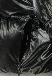 Sezione di giacca a vento nera con texture lucida e liscia. Presenta una tasca con zip e dettagli cuciti in un design compatto e voluminoso.
