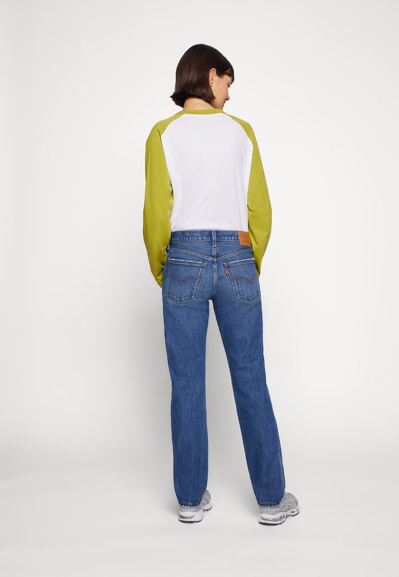 Camiseta de manga larga con cuerpo blanco y mangas verdes, combinada con vaqueros de pierna recta azules. Características destacadas incluyen una etiqueta de cuero marrón en la cinturilla.