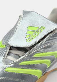 Srebrna atletska cipela s teksturiranim završetkom, neonsko zeleni detalji i upečatljivi preklop preko vezica. Sadrži Adidas brendiranje.
