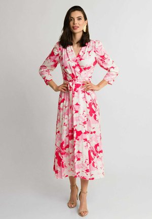 Kobieta w różowej, czerwonej i białej kwiecistej sukience na zakładkę z rękawami o długości do łokcia i beżowych sandałach na wysokim obcasie, stojąca z rękami na biodrach.