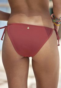 Bas de bikini marron avec liens sur les côtés, présentant une légère brillance et des détails cousus. Comprend un petit accent de logo près de la taille.