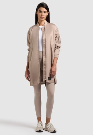 Woman wearing beige long bomber jacket, beige leggings, white crop top, and beige sneakers standing against plain background.