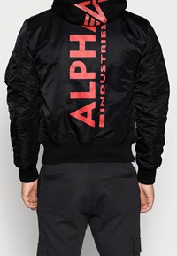 Giubbotto bomber nero con stampa rossa "ALPHA INDUSTRIES" sulla schiena, polsini e orlo a coste. Presenta una texture liscia e lucida.