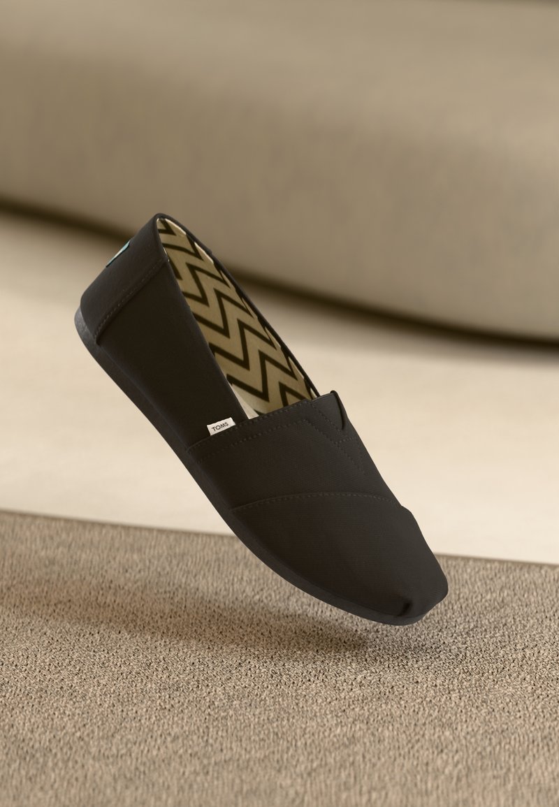 Chaussure slip-on noire en tissu texturé avec couture en angle. Intérieur avec motif en zigzag beige et marron. Conception légère et flexible.