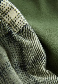 Olajzöld szövet mellett mintás textil világoskrémpöttyökkel és fekete pöttyökkel, változatos textúrákat és árnyalatokat bemutatva.