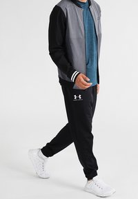 Person, der eine schwarze und graue Jacke, ein blaues Hemd, schwarze Under Armour-Sweatpants und weiße Sportschuhe trägt, geht vor einem einfachen Hintergrund.