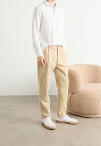 Osoba ubrana w białą koszulkę polo z długim rękawem, beżowe spodnie i białe trampki stojąca obok beżowego fotela o teksturowanej powierzchni na betonowej podłodze.