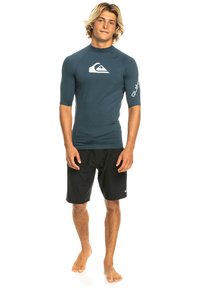 Men's dark blue short-sleeve rash guard with logo, paired with black board shorts. Fabric appears smooth and fitted, ideal for water activities.