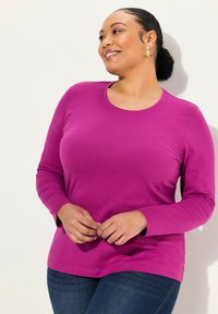 Langärmliges, runden Ausschnitt Shirt aus leuchtend magentafarbenem Baumwollstoff mit glatter Textur und taillierter Silhouette, kombiniert mit dunkelblauen Jeans.
