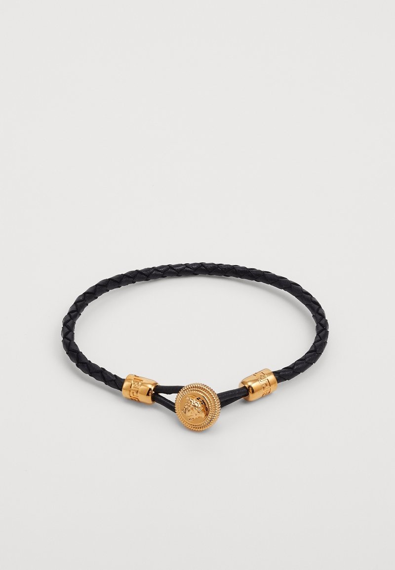 Bracelet en cuir tressé noir avec médaillon central tête de Méduse dorée et embouts cylindriques sur fond clair.