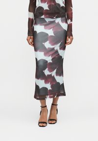 Γυναίκα που φοράει διάφανο, στενό σετ από φούστα και τοπ με μεγάλο μωβ λουλουδένιο σχέδιο, σε συνδυασμό με μαύρα πέδιλα με λουράκι στον αστράγαλο και ανοιχτό δάκτυλο.