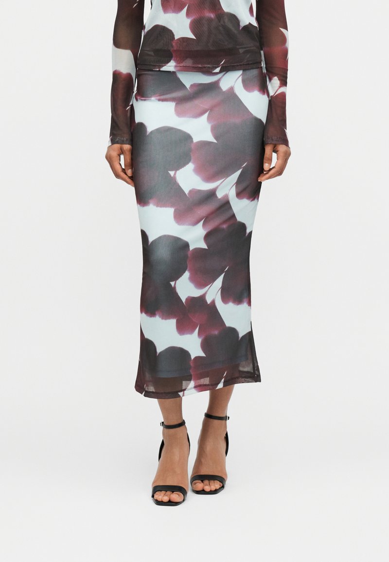 Γυναίκα που φοράει διάφανο, στενό σετ από φούστα και τοπ με μεγάλο μωβ λουλουδένιο σχέδιο, σε συνδυασμό με μαύρα πέδιλα με λουράκι στον αστράγαλο και ανοιχτό δάκτυλο.