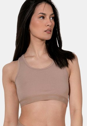 Donna con capelli scuri e lisci che indossa un reggiseno sportivo beige, che guarda di lato su uno sfondo chiaro e semplice.