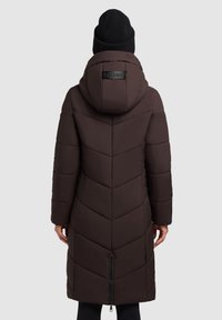 Brauner gesteppter Mantel mit Kapuze, Chevron-Muster, Reißverschluss und einem Logo-Akzent auf der Rückseite. Kombiniert mit einer schwarzen Strickmütze.