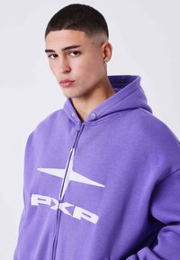 Sudadera de cremallera morada hecha de material suave, con un gran logotipo blanco y capucha. Incluye botones a presión en el cuello.