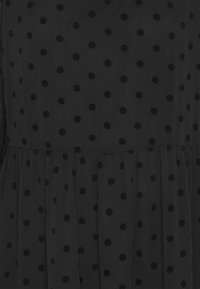 Mustat vaatteet, joissa on läpinäkyvää kangasta ja mustia pilkkuja. Ominaisuuksiin kuuluu kerätty vyötärö ja kevyt, liehuva rakenne.