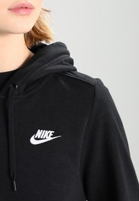 Close-up of a person wearing a black Nike hoodie with a white logo on the chest, showing part of the face and shoulder.
