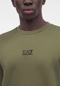 Крупен план на човек, носещ маслиненозелена риза с черно лого EA7 Emporio Armani на гърдите, на едноцветен фон.