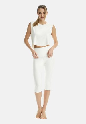 Donna in piedi a piedi nudi che indossa un top corto bianco senza maniche e leggings capri bianchi a vita alta su sfondo semplice.