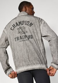 Szürke sportzakó, textúrált felülettel, hátulján vastag fekete betűkkel nyomtatva: "Champion XXL Training Academy".