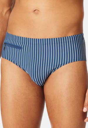 Man wearing blue swim briefs with thin white vertical stripes and a small zippered pocket on the left side.