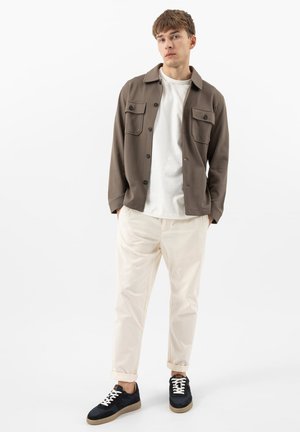 Jeune homme debout, les mains dans les poches, portant une veste marron boutonnée, un t-shirt blanc, un pantalon crème et des baskets noires sur fond blanc.