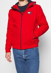 Rød hættejakke med lynlåslukning, med et logo patch på brystet, sidelommer og ribbede manchetter. Bæret over en navy sweater.