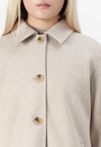 Beige wool coat with large marbled buttons worn over a knitted high-neck sweater, shown from the neck to mid-torso on a person.