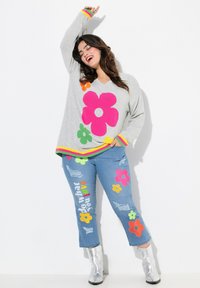 Pull-over gris clair en V avec des motifs de fleurs roses, oranges et vertes. Accompagné d'un jean en denim bleu avec des découpes et des accents multicolores.