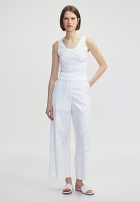 Weißes besticktes Tanktop kombiniert mit weißen weit geschnittenen Hosen mit floralem Ausschnitt. Das Model trägt weiße Sandalen mit geflochtenen Details.