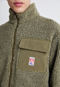 Giacca in pile verde oliva fuzzy con colletto alto, chiusura a zip e una grande tasca frontale sul petto con etichetta Wrangler.