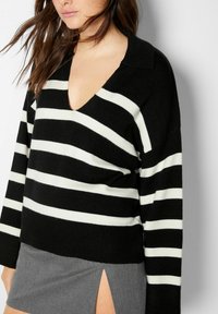 Femme portant un pull à col en V rayé noir et blanc et une jupe grise avec une fente haute, cheveux libres sur les épaules.