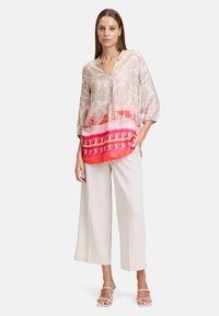 Langarmbluse in hellbeige mit Floralprint, mit einem rosa Farbverlauf am Saum und dekorativer Bordüre, kombiniert mit weiten weißen Hosen.