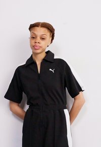 Svart kortermet jumpsuit med hvite sidestriper, glidelås foran og Puma-logo på brystet. Laget av glatt stoff. Tettsittende i midjen.