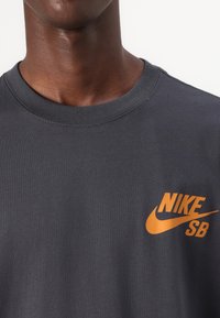 Mørkegrå T-shirt lavet af bomuld, med et orange Nike SB-logo på venstre bryst. Halsudskæringen er i klassisk crew-stil.
