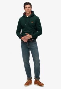 Groene katoenen hoodie met een logo, gelaagd over een rood geruit overhemd, gecombineerd met blauwe denim jeans en bruine veterschoenen.