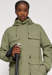 Grüne, kapuzenjacke aus glattem Material mit zwei Fronttaschen und verstellbaren Kordeln. Eine Person hält einen schwarzen Wanderstock.