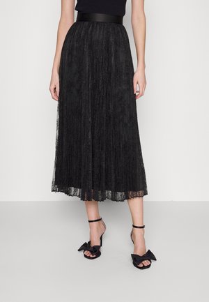 Femme portant une jupe midi plissée en dentelle noire avec une large ceinture et des talons ouverts noirs avec des détails en nœud.