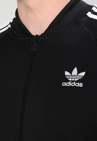 Černá zipová bunda vyrobená z hladkého materiálu, s bílým vyšitým logem Adidas a ikonickými třemi pruhy na rukávech.