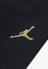 Un material textil negru cu o textură netedă, înfățișează un logo Jumpman brodat cu fir aurit, într-o poziție dinamică, subliniind mișcarea atletică.