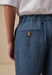 Blauwe linnen broek met een gestructureerde afwerking, voorzien van een achterzak en een enkele bruine knop die de tailleband accentueert.
