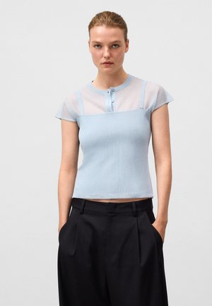 Haut en tricot bleu clair à manches courtes, avec des détails en maille aux épaules et une patte de boutonnage. Associé à un pantalon noir taille haute.
