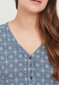 Blouse à motifs bleus avec un décolleté en V, quatre boutons sur le devant et des motifs floraux texturés dans différentes nuances de bleu et blanc.